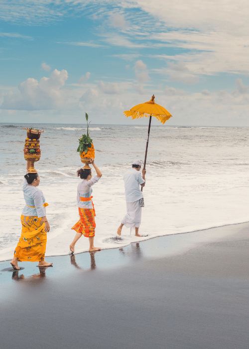 Two girls and one man in traditional Balinese dress walking on the beach for a religious ceremony, honoring cultural rituals by the sea.