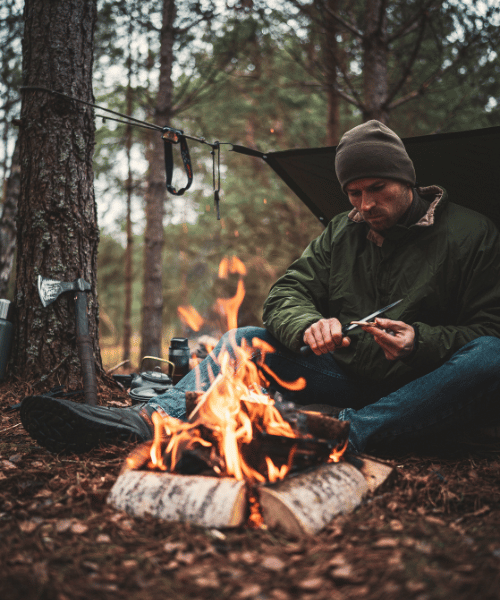 A man cutting wood in front of a campfire during jungle camping, preparing for the night in a peaceful forest setting