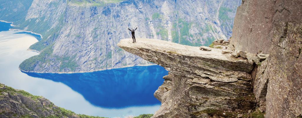 A solo hiker standing triumphantly on Trolltunga rock ledge in Norway, overlooking vast fjords and dramatic mountain scenery