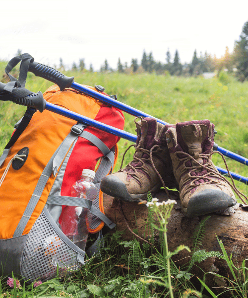 Various hiking and camping tools laid out neatly on green grass, including backpack, poles, boots, and navigation gear