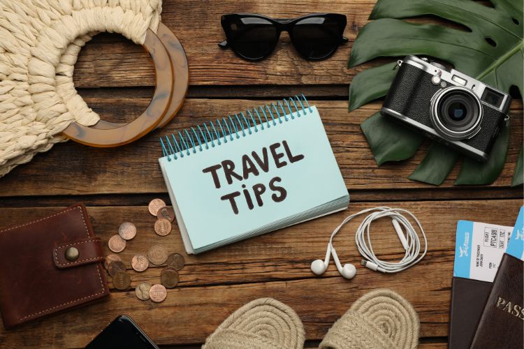 A flat-lay photograph on a wooden surface featuring travel essentials like a camera, sunglasses, a wallet with coins, and a notebook that reads "TRAVEL TIPS.