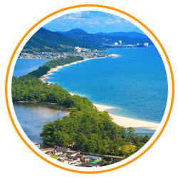 An aerial circular view of the Amanohashidate sandbar in Kyoto Prefecture, featuring a long, pine tree-covered path stretching across a blue bay.