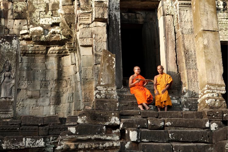 Two Buddhist monks in traditional orange robes sitting on the stone steps of an ancient Khmer temple in Cambodia.