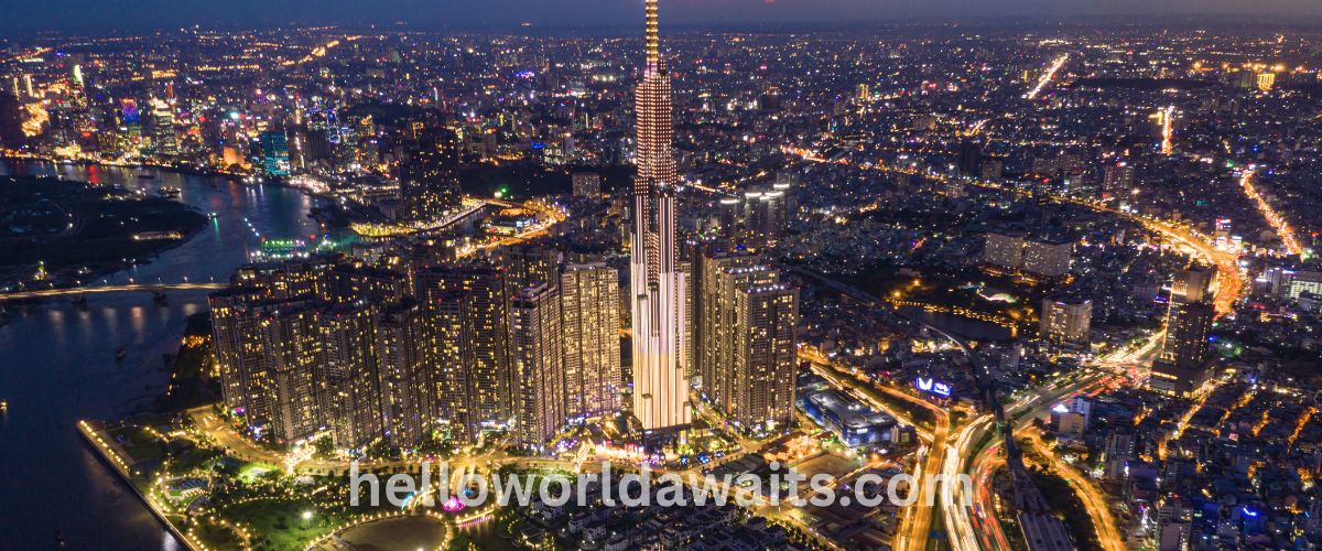 A stunning night aerial view of the Ho Chi Minh City skyline, dominated by the illuminated Landmark 81 skyscraper in the center. The city is alive with golden lights from dense clusters of high-rise apartment buildings and winding highways filled with the light trails of traffic. A dark river curves around the base of the city on the left side, reflecting the city lights. The watermark "helloworldawaits.com" is visible at the bottom.