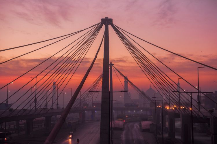 A vibrant sunset over the cable-stayed Azadi Interchange in Lahore, with the silhouetted minarets and domes of the Badshahi Mosque visible in the hazy background