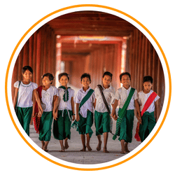 A group of smiling Myanmar school children walking together through a traditional wooden temple corridor, wearing green and white uniforms with colorful sashes.