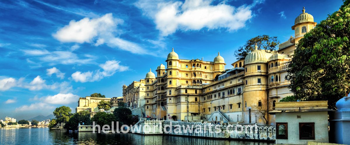 A wide shot of a grand, multi-story yellow palace with many domes and windows, situated directly on the edge of a body of water. The sky is bright blue with scattered white clouds, and lush green trees are visible to the right.