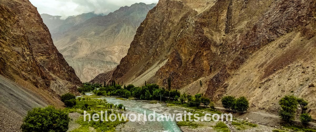 A winding turquoise river flowing through a deep valley surrounded by towering, rugged brown mountains and a strip of lush green trees under a cloudy sky
