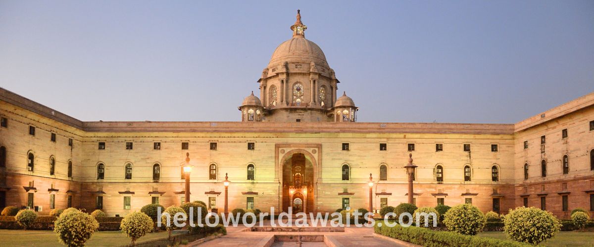 A symmetrical view of the Rashtrapati Bhavan in New Delhi at dusk. The large sandstone building features a prominent central dome and long wings with rows of windows, fronted by a manicured garden with spherical bushes and glowing street lamps.