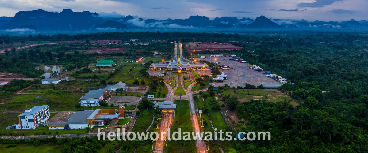 High-angle aerial view of a brightly lit border crossing facility at dusk, surrounded by dark forests and mist-covered mountains in Laos.