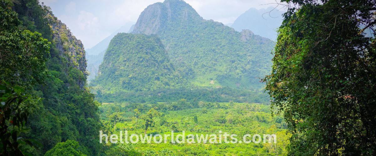 Panoramic view of a lush green valley in Vang Vieng, Laos, featuring towering limestone karst mountains covered in tropical vegetation under a bright sky, framed by dense jungle foliage in the foreground.