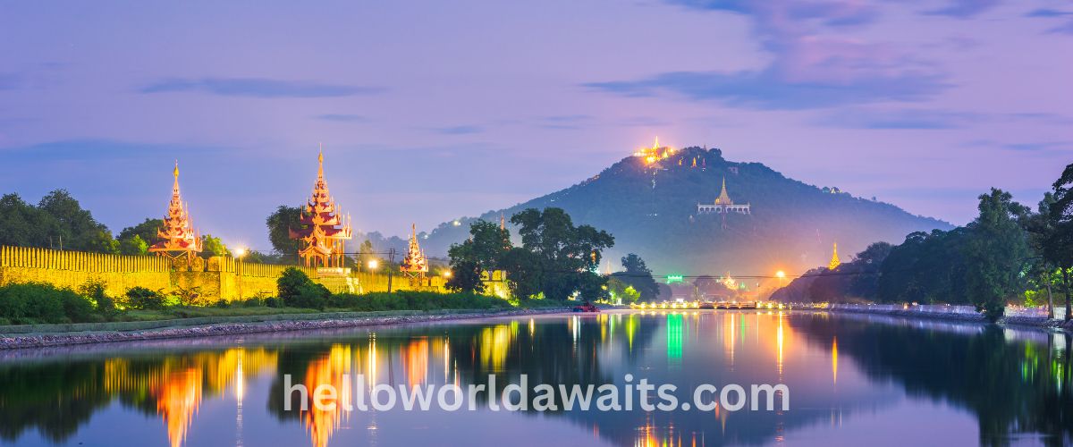 A scenic twilight view of the Mandalay Palace moat in Myanmar, reflecting illuminated golden spires and city lights in the calm water, with the lit-up temples of Mandalay Hill visible in the background against a purple sky.