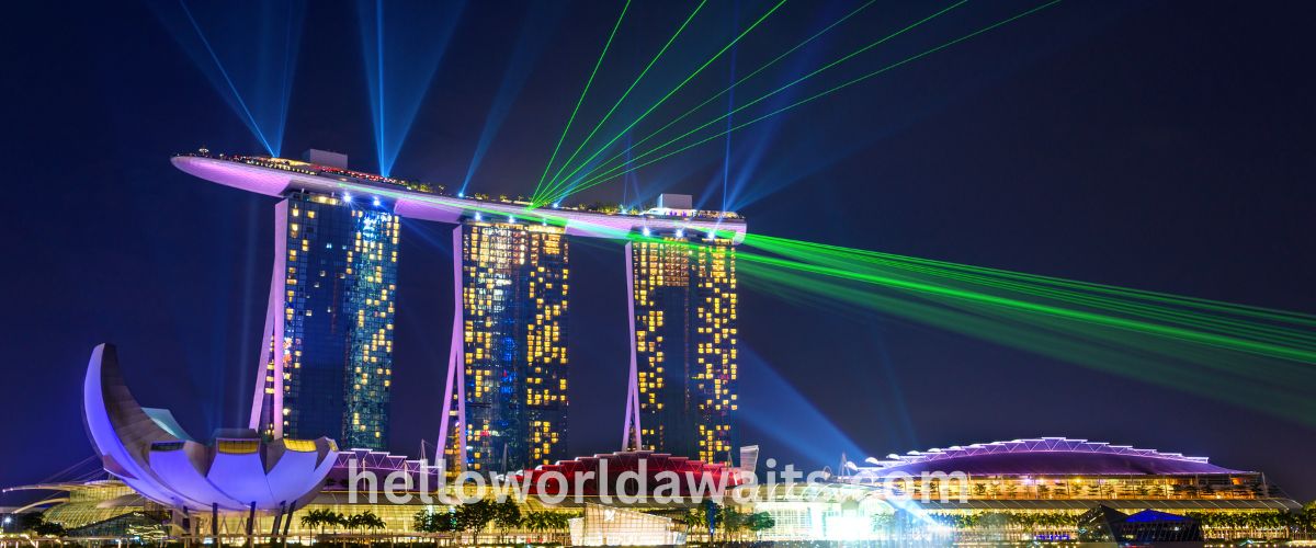 Night view of Marina Bay Sands in Singapore during the Spectra light and water show, featuring blue and green laser beams shooting from the Sands SkyPark across the dark sky.