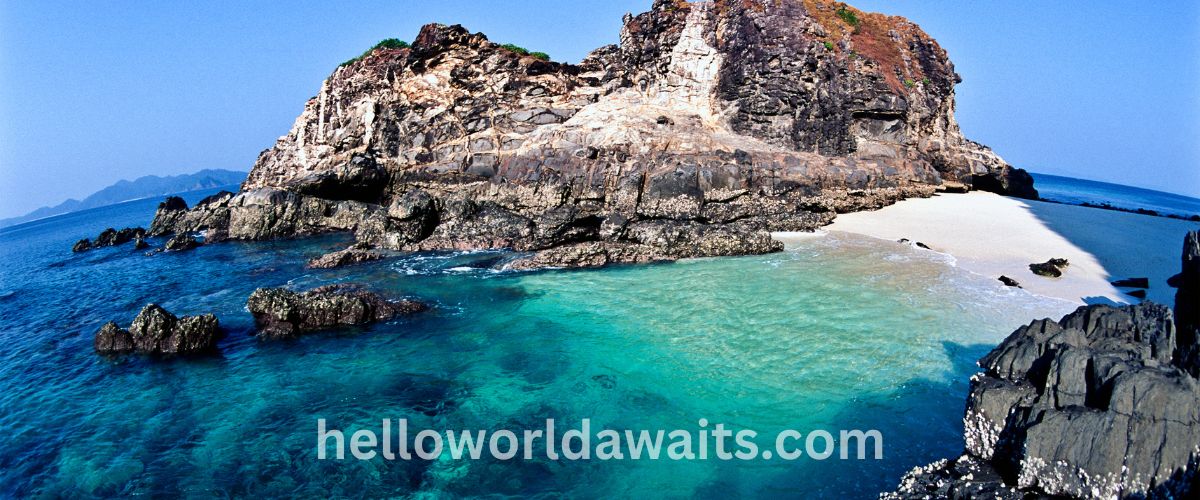 A panoramic view of a rocky island in Myanmar featuring crystal clear turquoise water, a small white sand beach, and dark jagged rock formations under a bright blue sky.