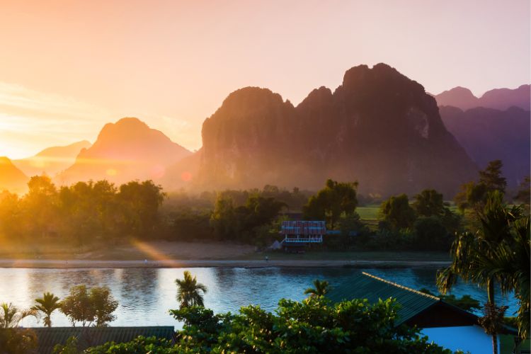 A warm, orange sunset over layered, misty mountain peaks and a river valley in Luang Prabang, Laos, with the bright sun low in the sky casting deep shadows over the rugged terrain.