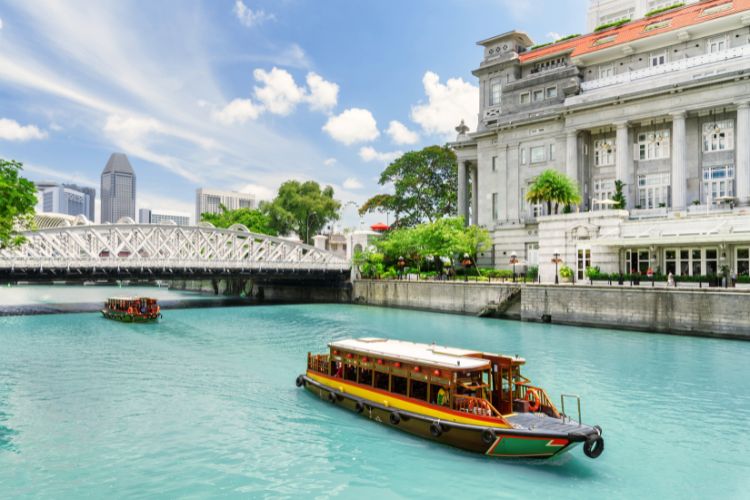 A traditional bumboat cruising along the Singapore River with the historic Fullerton Hotel and Anderson Bridge in the background.