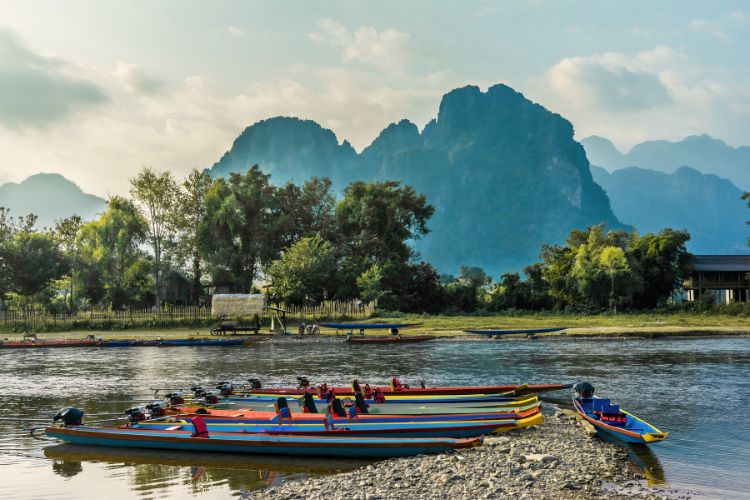 A row of colorful, long-tail motorized boats docked on a rocky shore of the Nam Song River in Vang Vieng, Laos, with lush green trees and dramatic limestone karst mountains under a hazy blue sky in the background.