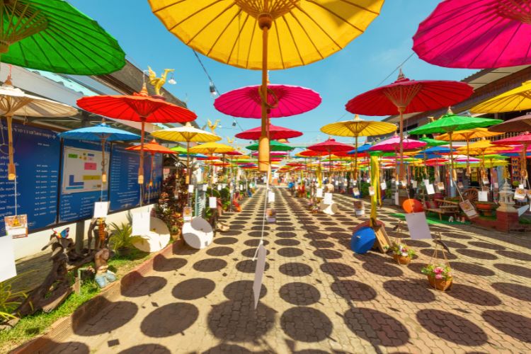A vibrant, low-angle shot of colorful, hand-painted Thai paper umbrellas hanging from a bamboo structure against a bright blue sky.