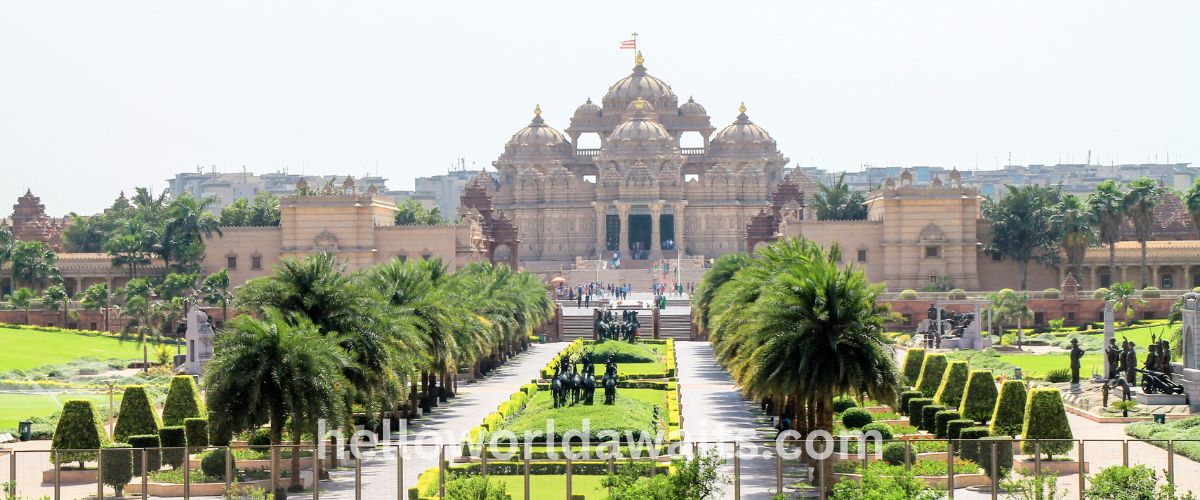 Wide shot of the grand Akshardham Temple in New Delhi, featuring intricate sandstone carvings, multiple domes, and a central golden pinnacle. The foreground shows lush green gardens, rows of palm trees, and a long walkway leading to the temple entrance under a bright sky