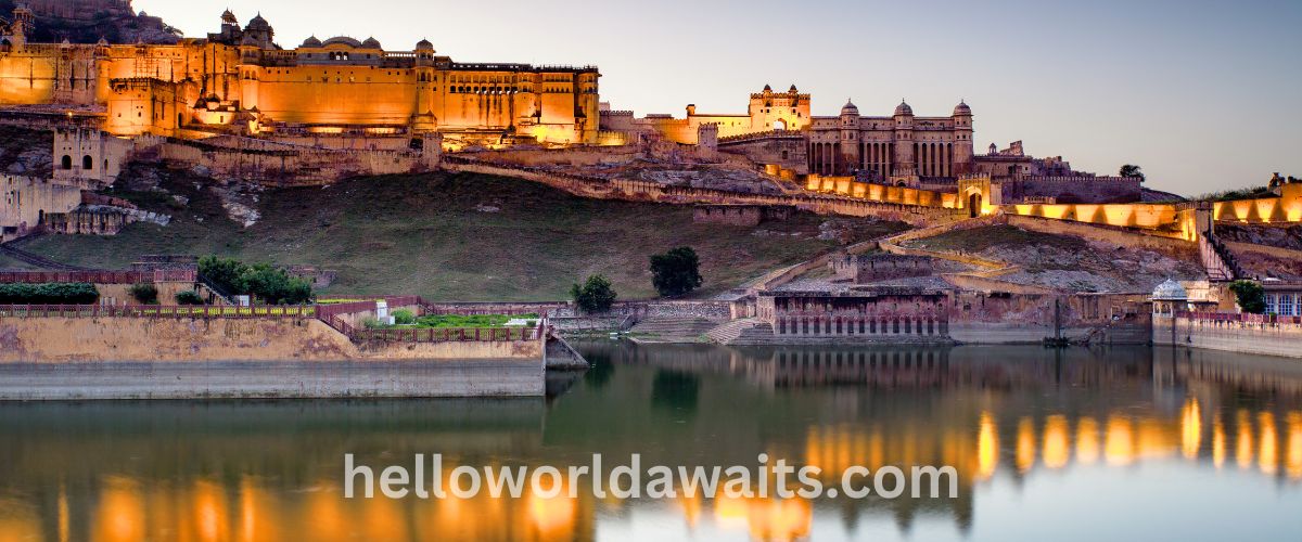 A wide panoramic view of the Amer Fort in Jaipur at twilight. The massive sandstone palace complex is illuminated with warm golden lights, reflecting beautifully in the calm waters of Maota Lake in the foreground.