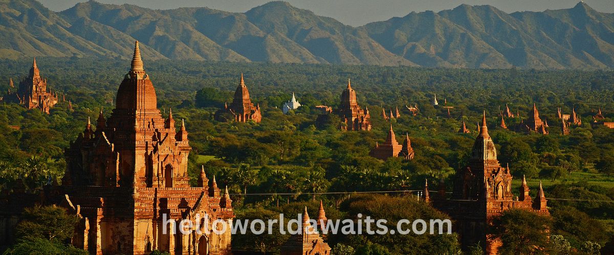 A vast landscape view of the Bagan plains in Myanmar, filled with numerous ancient brick temples and pagodas nestled among green trees, with a dramatic mountain range in the background.