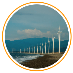 A scenic view of the Bangui Wind Farm in Ilocos Norte, Philippines, showing a long row of tall white wind turbines lined up along a dark pebble beach with ocean waves crashing and blue mountains in the background.