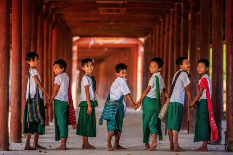 A group of seven smiling Burmese schoolchildren holding hands and walking through a traditional wooden colonnade in Myanmar. They are wearing white shirts and green longyis, carrying woven traditional bags.