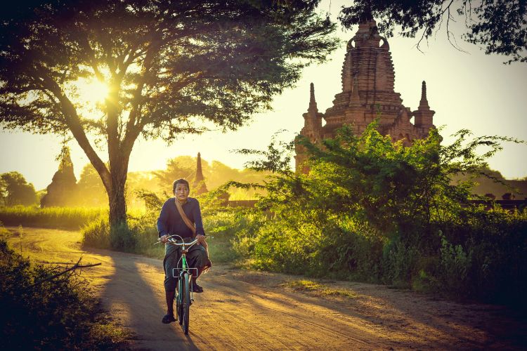 An oval-framed photograph of a man riding a bicycle down a dusty dirt path at sunset, with a large ancient brick Buddhist stupa and lush trees in the background.