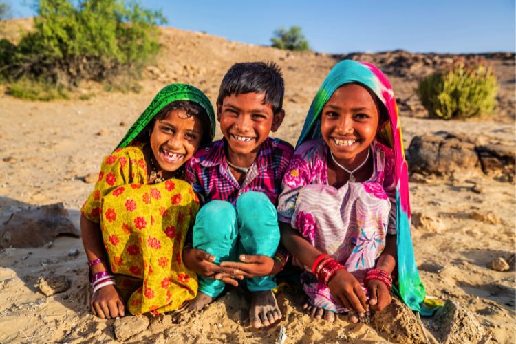 Three young children sitting together on sandy ground, smiling brightly at the camera. The two girls wear vibrant, patterned traditional dresses and colorful headscarves in green and pink.