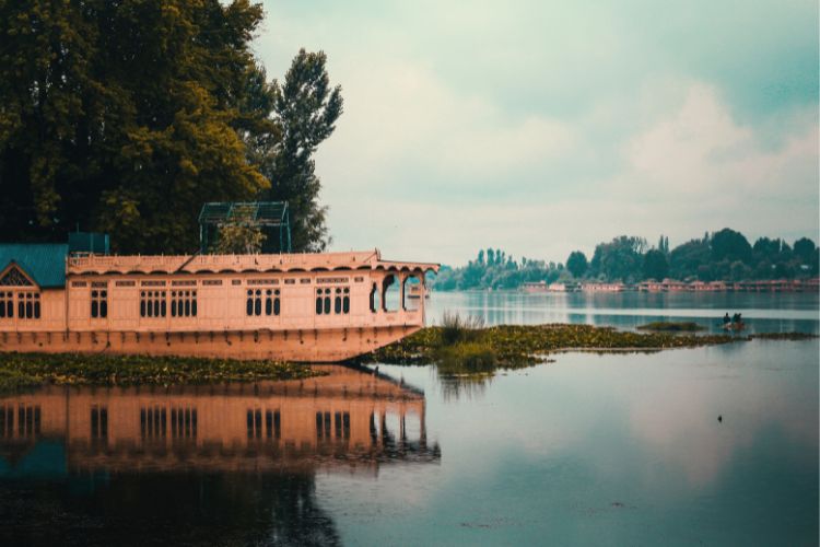 A traditional wooden houseboat reflected in the calm waters of Dal Lake, Srinagar, Kashmir, surrounded by lush greenery under a cloudy sky