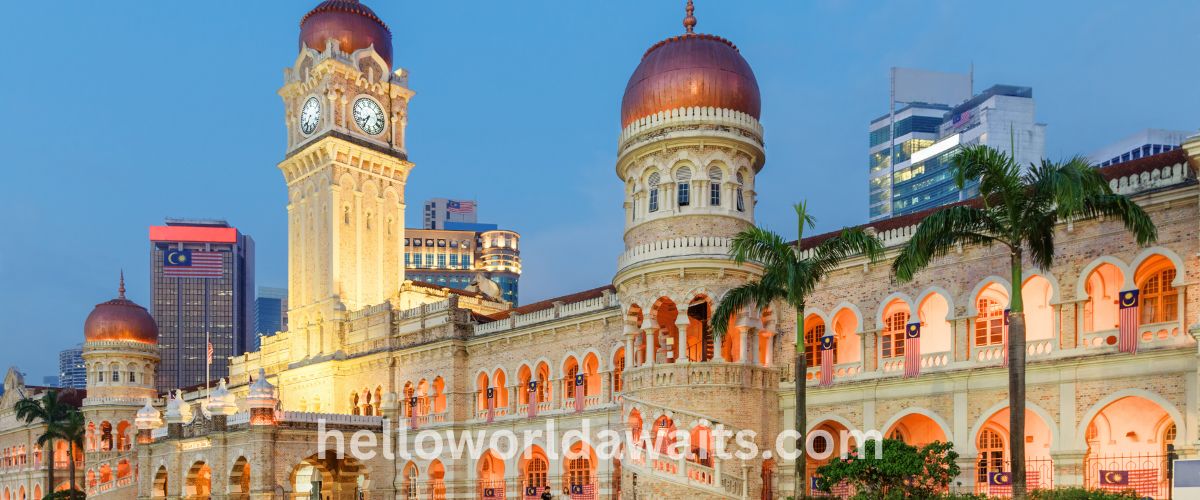 A twilight view of the Sultan Abdul Samad Building in Kuala Lumpur, Malaysia, illuminated with warm lights. The grand Moorish-style architecture features brick walls, arched windows, and copper-domed towers, including the prominent central clock tower. Malaysian flags hang from the arches, and modern city skyscrapers are visible in the background under a deep blue dusk sky. A watermark for "helloworldawaits.com" is centered at the bottom.
