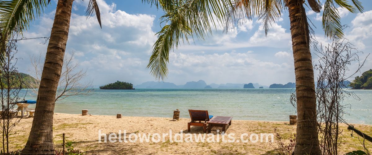 A peaceful tropical beach in Thailand featuring white sand, palm trees framing the view, and a lone wooden lounge chair looking out over turquoise water toward distant limestone islands.