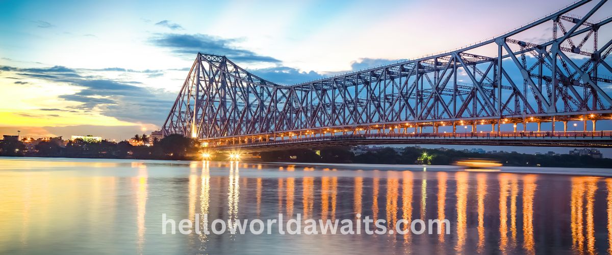 A long-exposure sunset view of the Howrah Bridge in Kolkata, a massive steel cantilever structure spanning a wide river. The bridge lights and the colors of the sunset reflect brightly on the calm water surface.