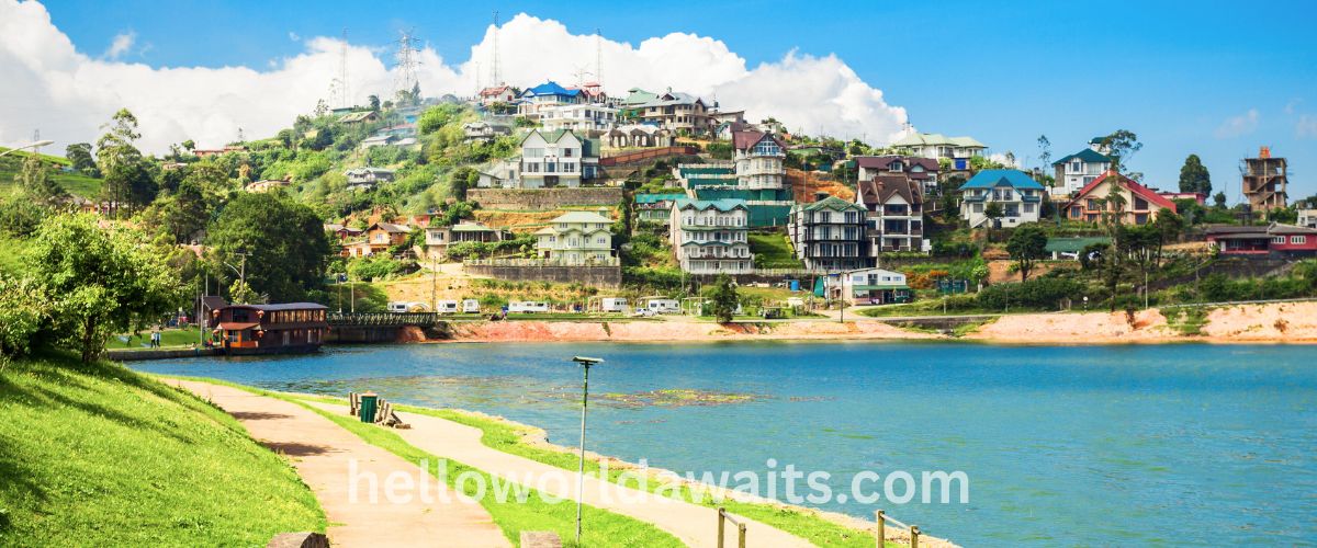 A scenic view of Gregory Lake in Nuwara Eliya, Sri Lanka, featuring colorful houses on a lush green hillside overlooking calm blue waters. A walking path and recreational boat are visible, capturing the charm of Sri Lanka's "Little England" hill country.