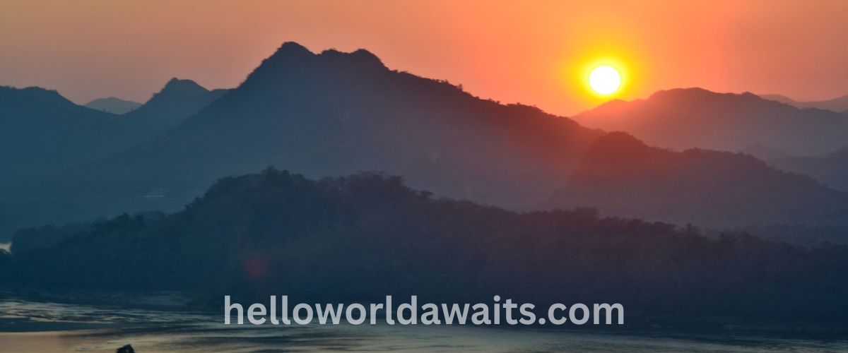 A warm sunset over a series of layered, misty mountain peaks in Luang Prabang, Laos, with the bright sun low in an orange sky casting deep shadows over the rugged terrain.