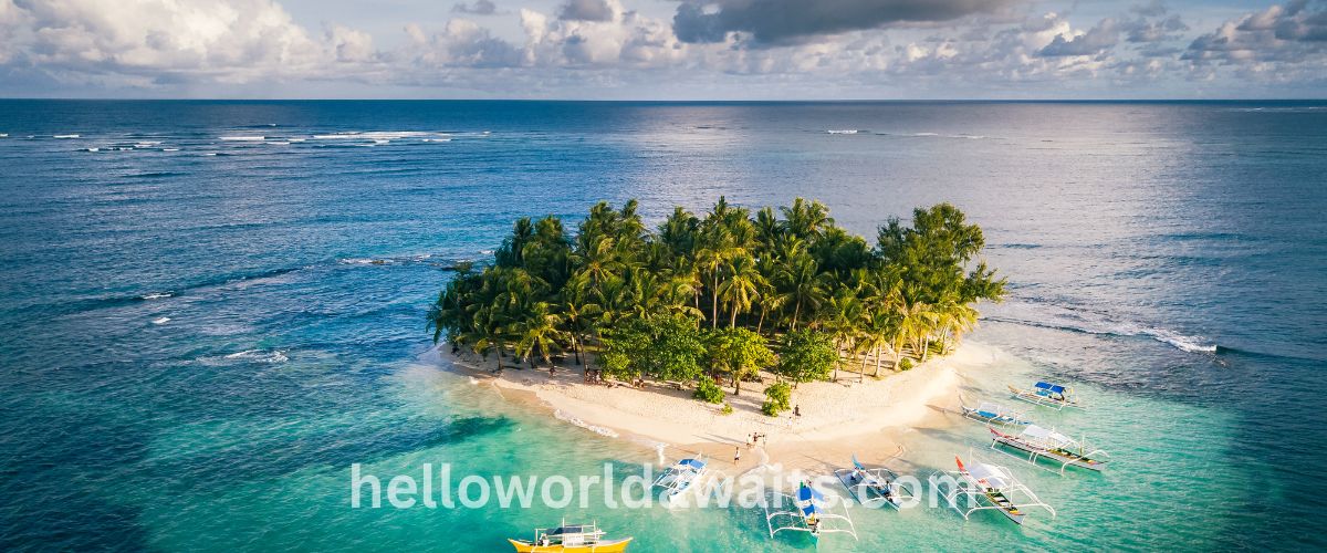 An aerial view of Guyam Island in Siargao, Philippines, showing a small tropical island with white sand and palm trees surrounded by clear turquoise water with several traditional outrigger boats (Bangkas) anchored at the shore.