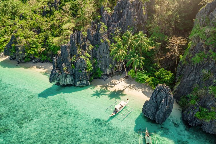 Aerial view of a traditional Bangka boat anchored in the turquoise waters of El Nido, Palawan, featuring towering limestone cliffs, white sand beaches, and lush tropical palm trees in the Philippines.