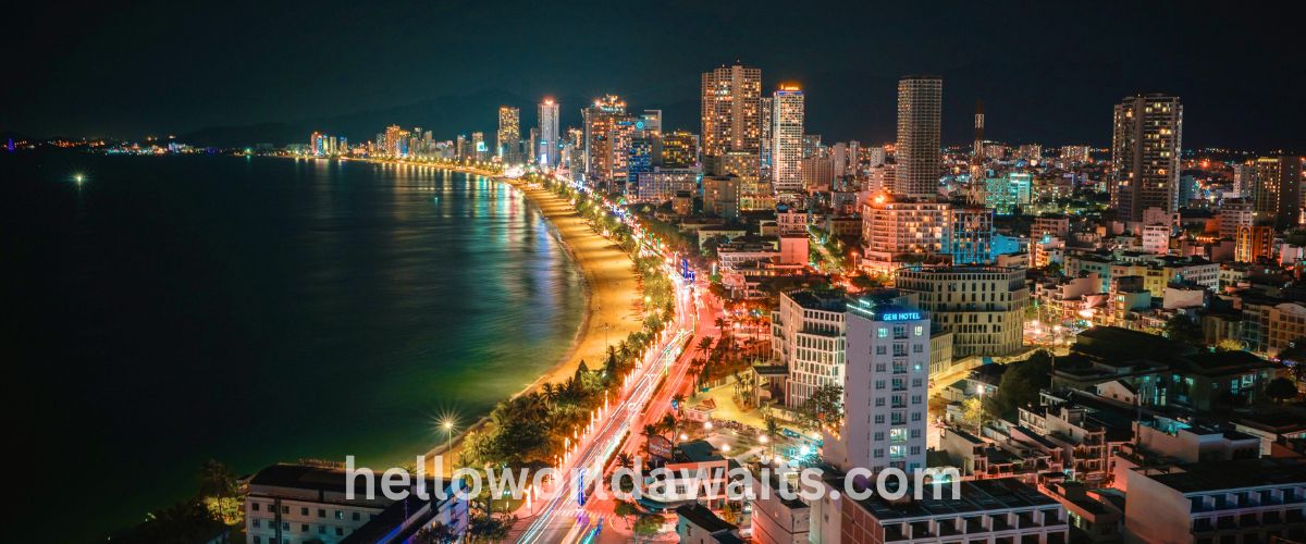 A vibrant night view of the Nha Trang city skyline in Vietnam, showing illuminated skyscrapers, a curved sandy beach, and glowing streetlights reflecting on the dark ocean water.