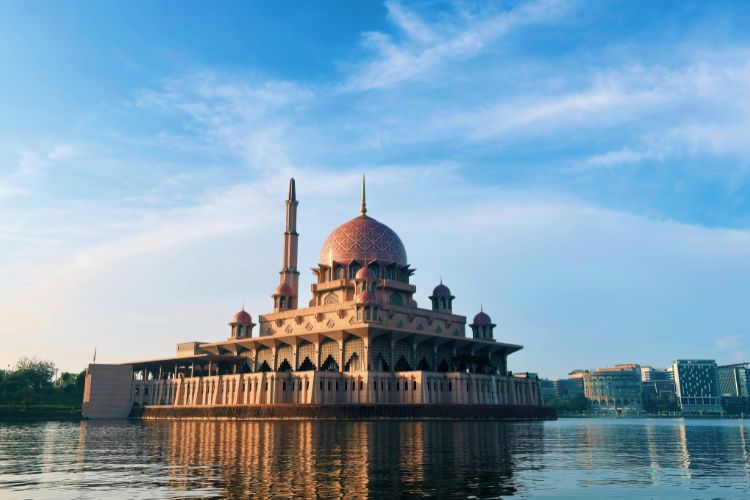 A wide-angle shot of the Putra Mosque in Putrajaya, Malaysia, featuring its iconic pink granite dome and tall minaret.