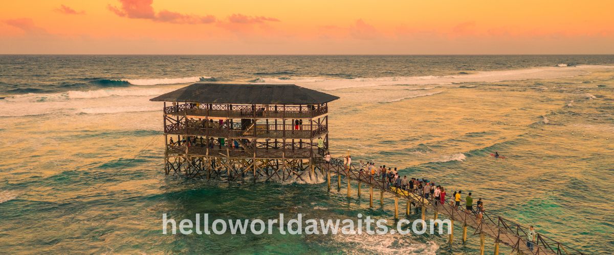 An aerial sunset view of the famous wooden Cloud 9 surfing boardwalk and three-story viewing deck in Siargao, Philippines, extending over the ocean with waves breaking nearby and people walking along the pier.