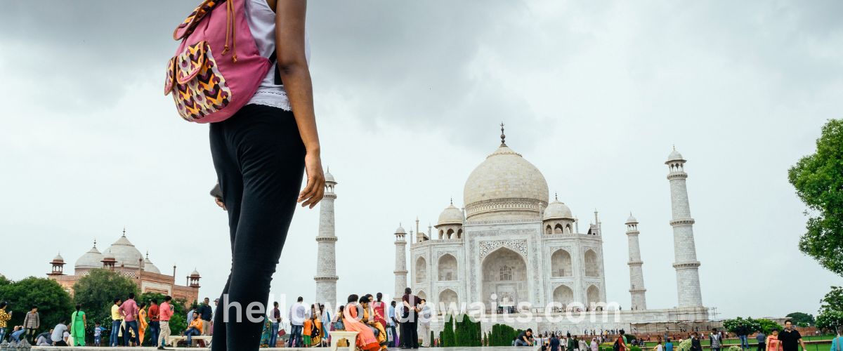 A traveler with a colorful pink and patterned backpack stands in the foreground, looking toward the white marble Taj Mahal in Agra, India. The scene shows the iconic central dome and minarets under an overcast sky with other tourists visiting the grounds.
