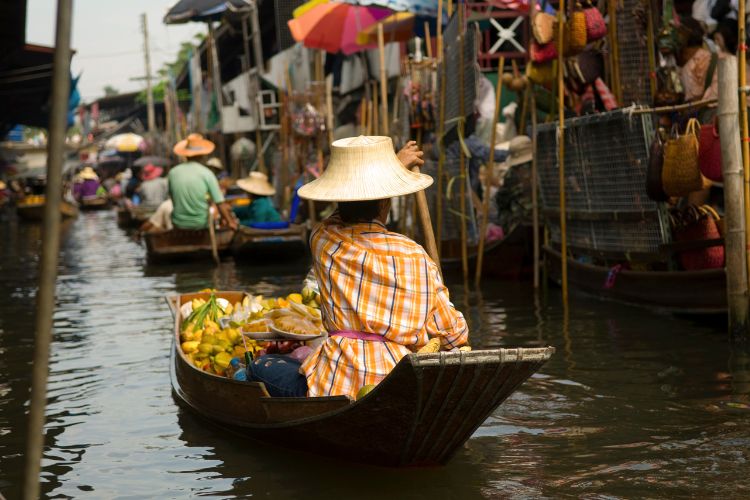 A vendor in a traditional straw hat paddles a wooden boat filled with fresh tropical fruits through a crowded, bustling floating market in Thailand.