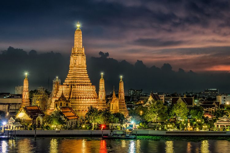 The illuminated Wat Arun temple (Temple of Dawn) at night, reflected in the Chao Phraya River under a dramatic sunset sky in Bangkok, Thailand.