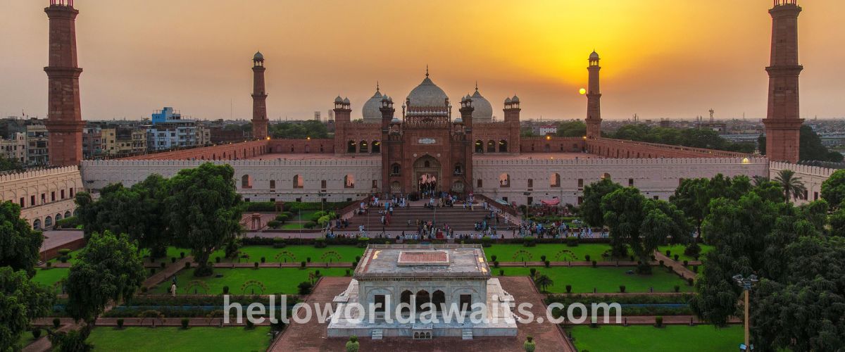 A wide aerial view of the historic Badshahi Mosque in Lahore at sunset, featuring its massive red sandstone courtyard, white marble domes, and the white marble Hazuri Bagh Baradari monument in the foreground garden.