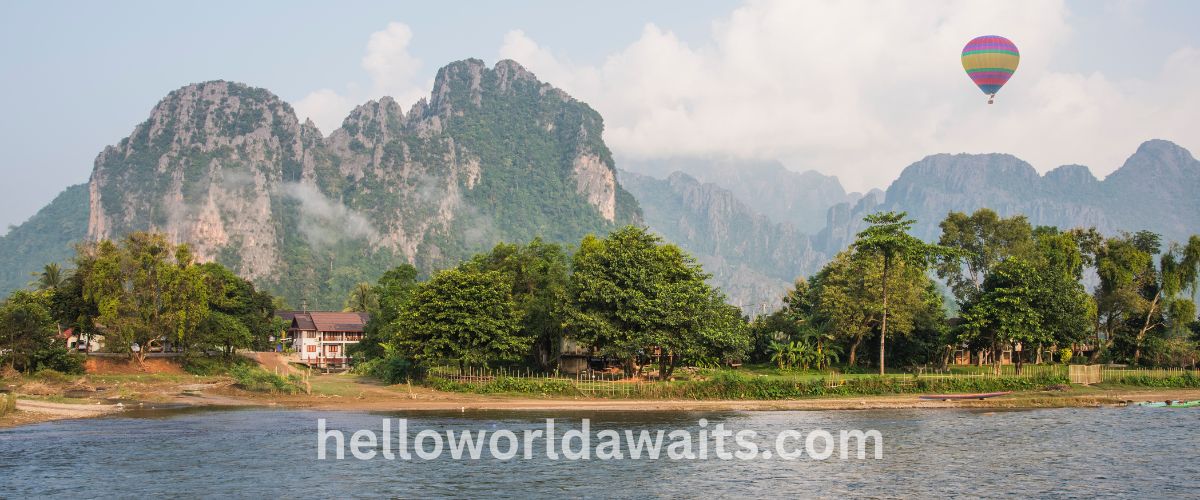 A colorful hot air balloon floats in a hazy sky above the Nam Song River and dramatic limestone karst mountains in Vang Vieng, Laos.