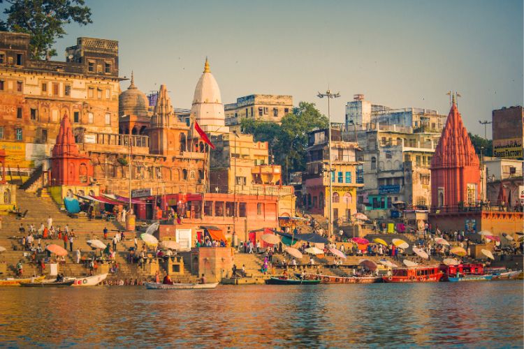 A vibrant view of the historic Dashashwamedh Ghat in Varanasi, India, featuring colorful ancient buildings, temples with red and white spires, and numerous boats docked along the banks of the Ganges River at sunset.