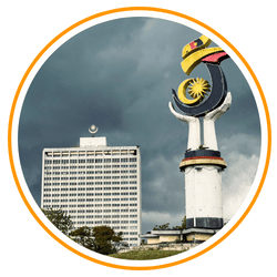 A circular-framed photo featuring the Malaysian Parliament House and a nearby monument against a dark, cloudy sky.