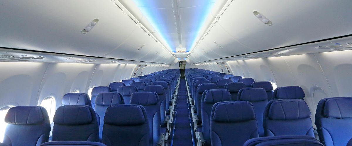 Empty airplane cabin interior with rows of blue seats and a center aisle, lit by soft blue mood lighting along the ceiling.