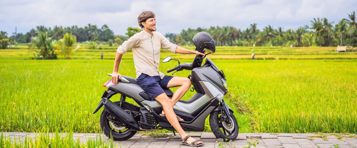 Tourist sitting on a scooter beside green rice fields, smiling while figuring out how to get to Bali and explore beyond the airport.