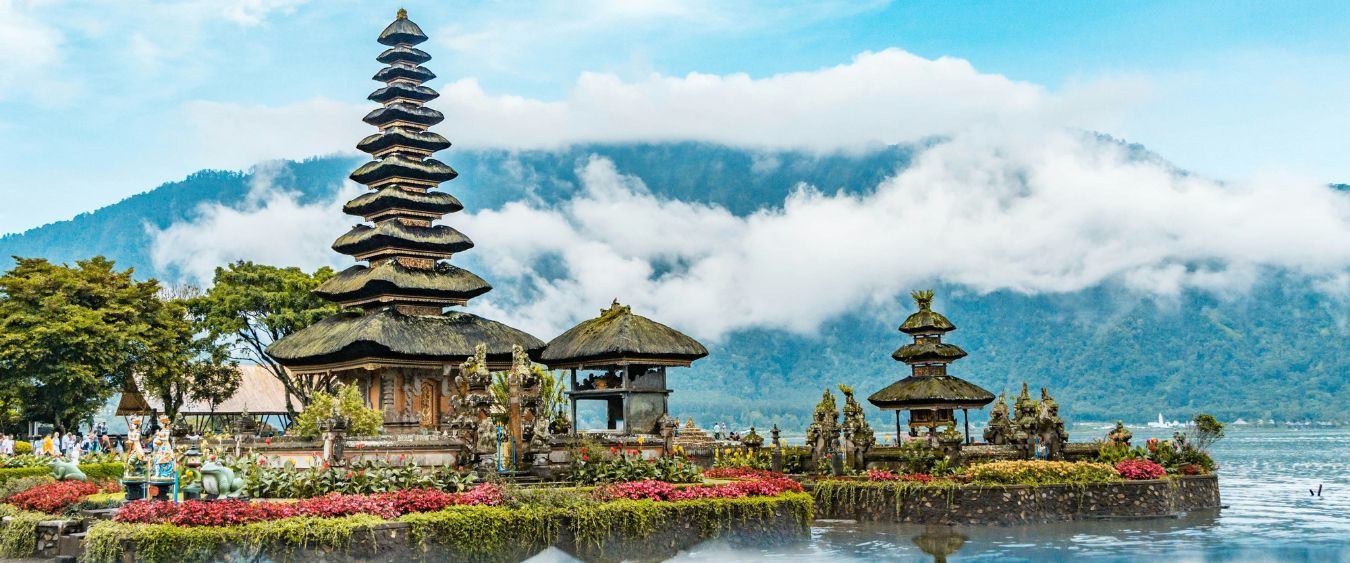 Pura Ulun Danu Bratan, a multi-tiered Balinese Hindu temple, sits on Lake Bratan in Bali with misty mountains in the background and colorful flowers along the shore.
